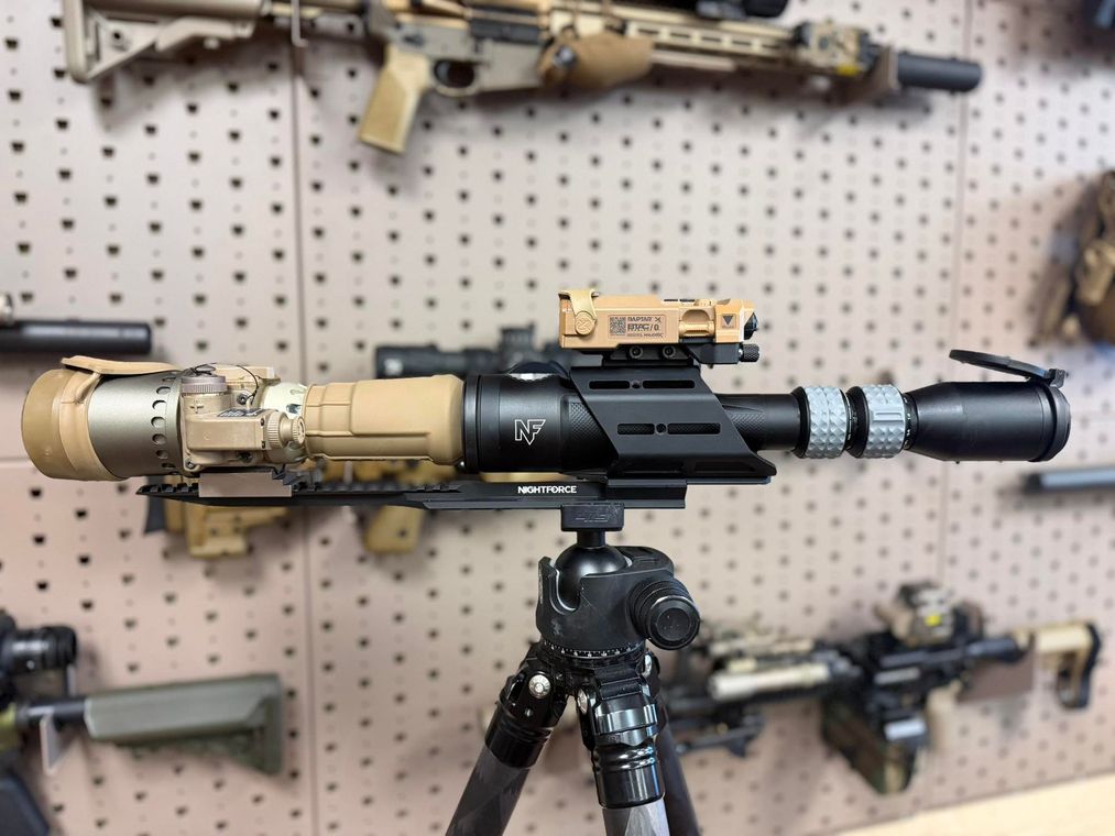 Rifle mounted on a tripod in a display room, with other firearms visible on a pegboard wall.