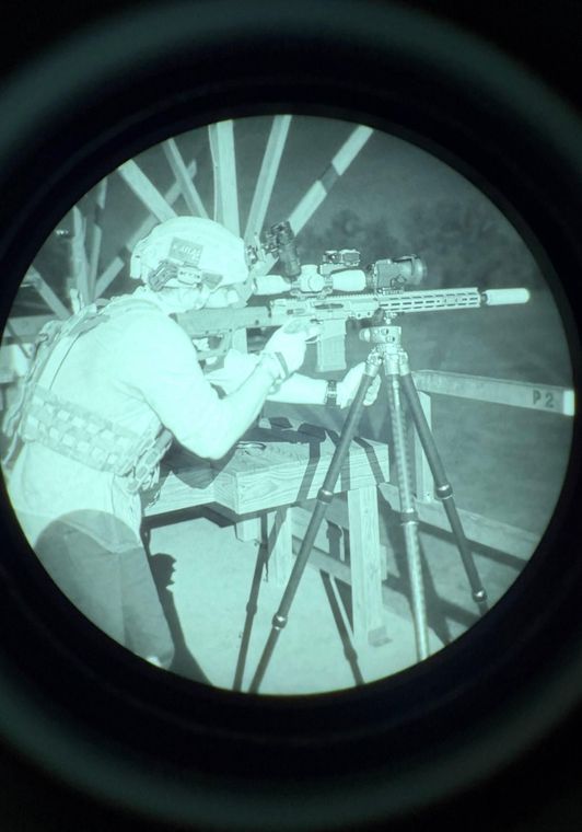 Night-vision view of a person aiming a rifle on a tripod at an outdoor shooting range.