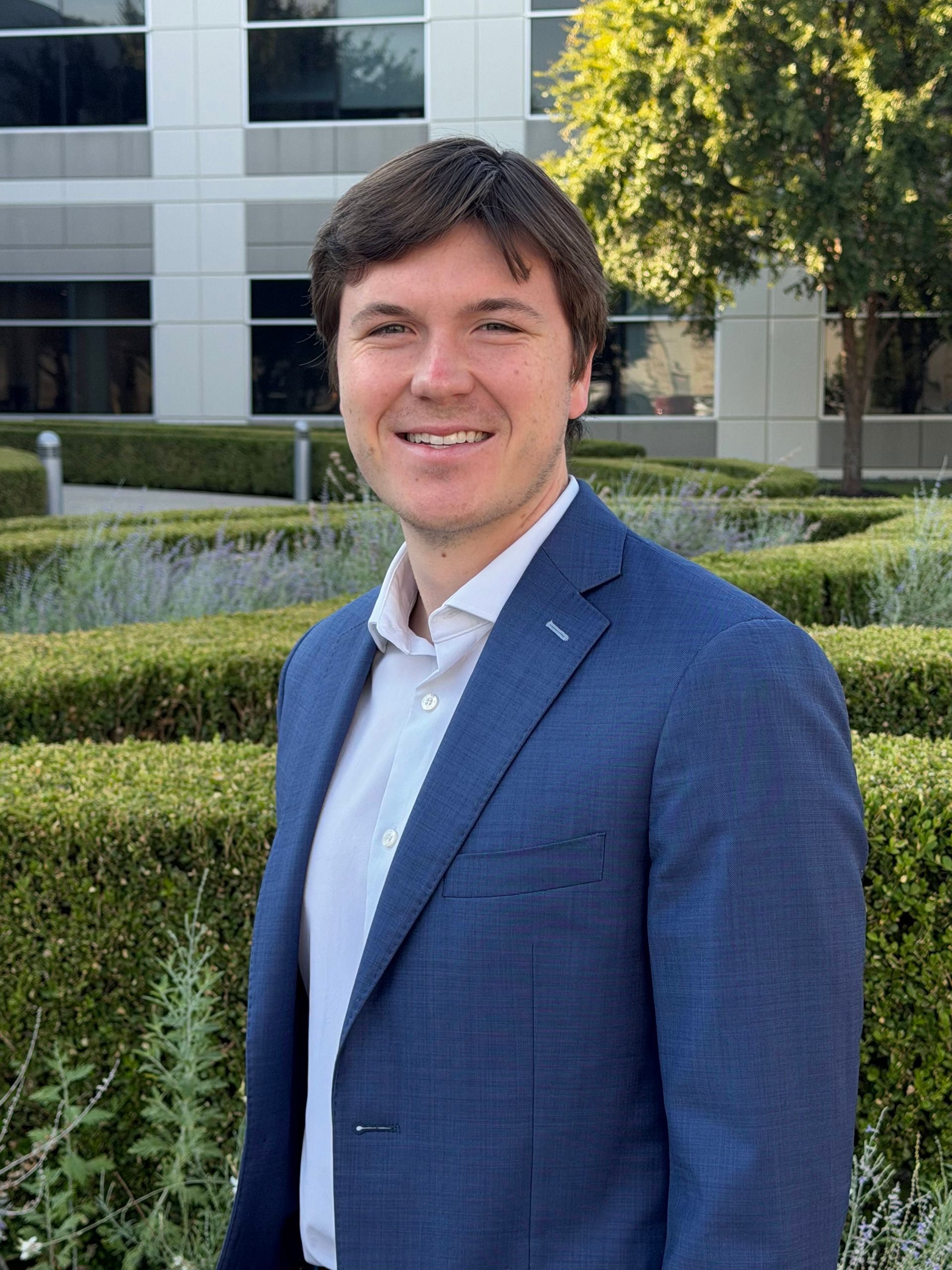 Man in a blue blazer smiling outdoors in front of an office building and hedges