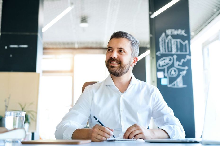 Man with a beard in a white shirt, smiles while holding a pen at a desk in an office.