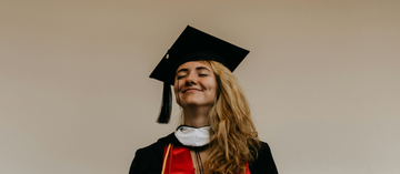 Person in graduation cap and gown smiles with eyes closed against a neutral background.