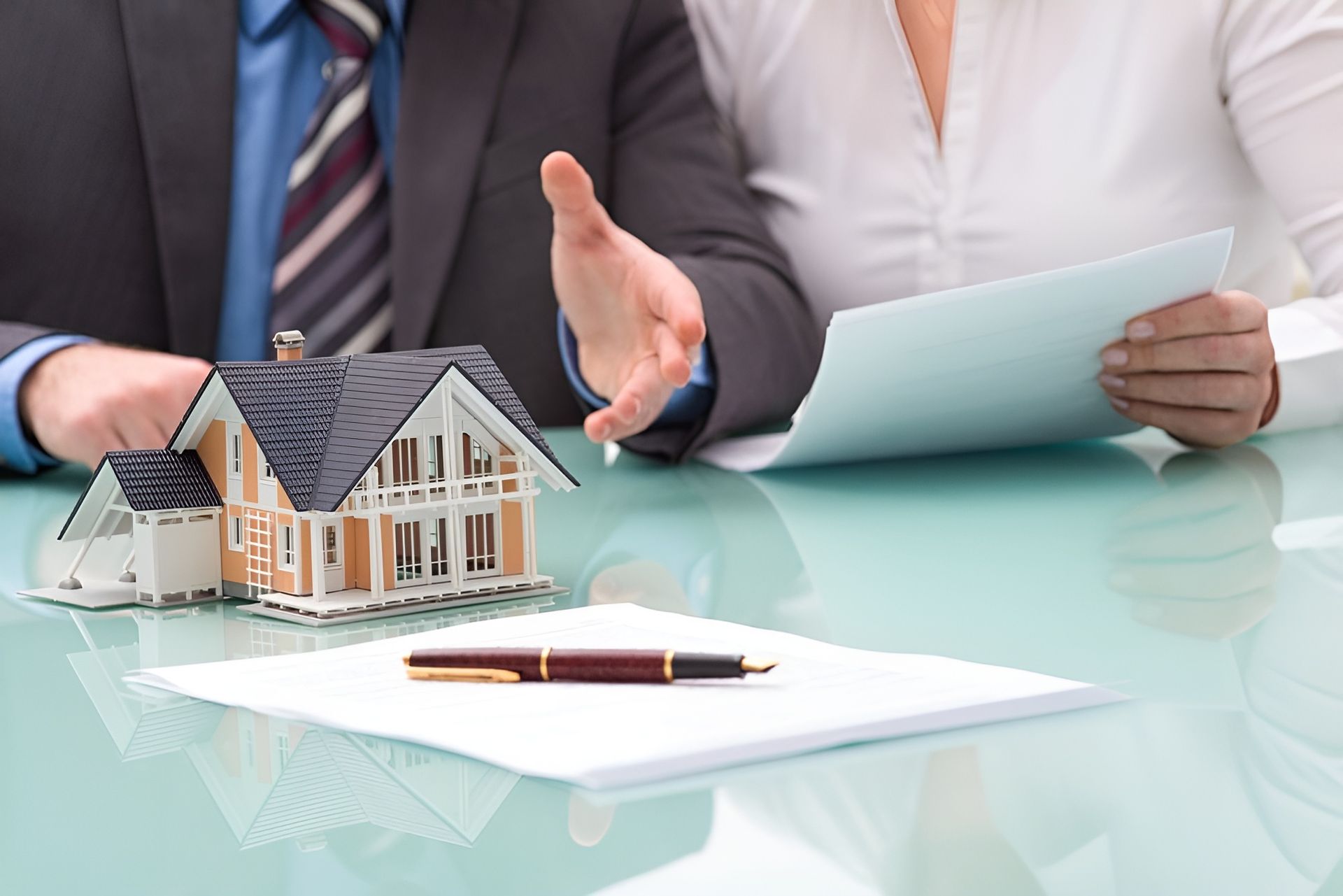 A real estate agent gestures toward a miniature house model on a table with a contract and a pen — Murphy Finance Solutions In Zuccoli, NT