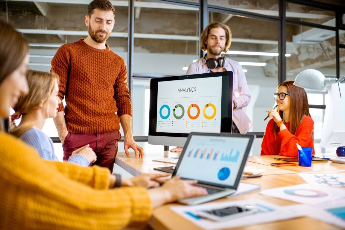Team meeting in a bright office, with a presenter at a screen and coworkers using laptops at a table