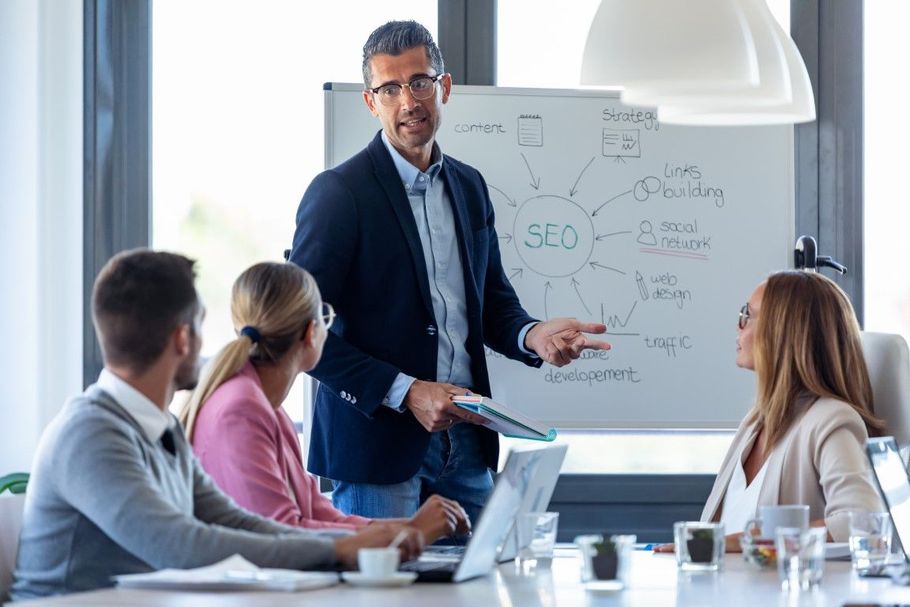 Business meeting with presenter at whiteboard speaking to seated colleagues in a bright conference room