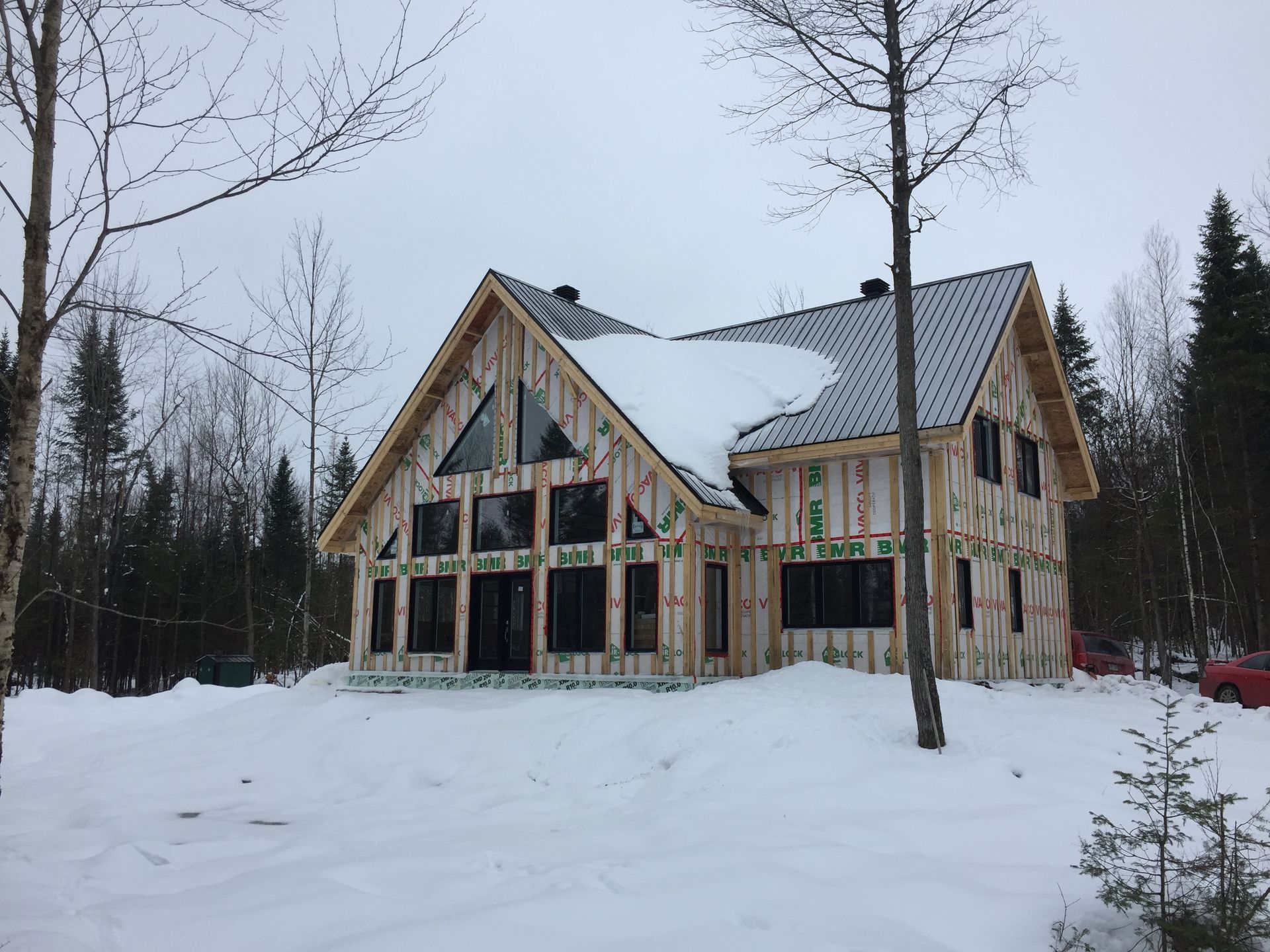 Maison en construction dans un paysage enneigé, charpente en bois, fenêtres sombres et neige sur le toit.