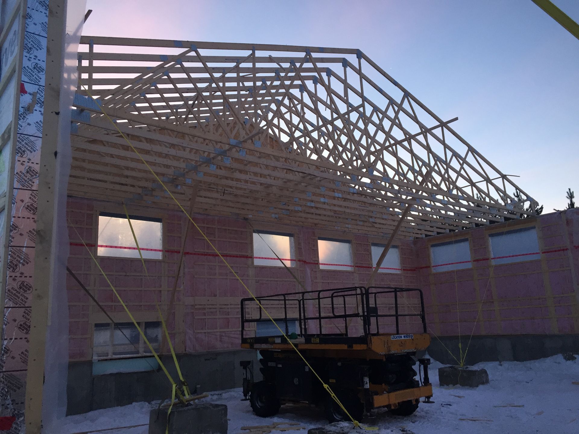 Chantier : installation de fermes de toit en bois sur un bâtiment en briques, soutenues par des câbles. Une nacelle élévatrice est visible devant.
