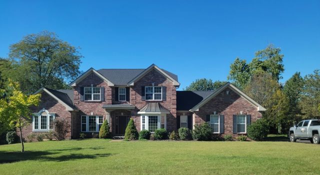 Large brick suburban house with a manicured lawn, blue sky, and a white truck parked at the right edge