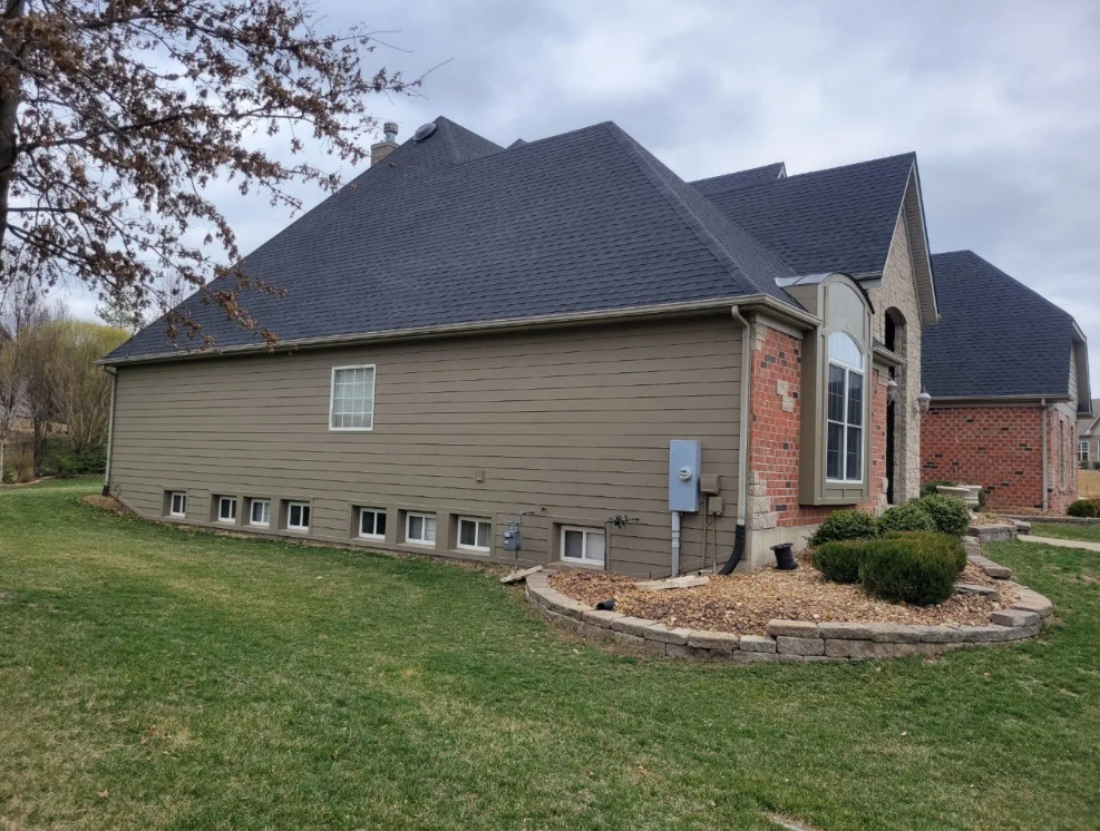 Suburban house exterior with tan siding, dark roof, brick entry, and green lawn