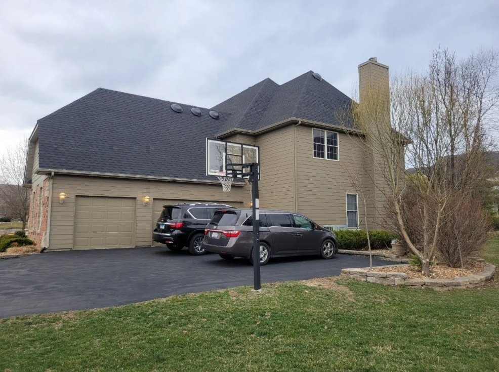 Suburban two-story house with dark roof, garage, driveway, and three parked cars under an overcast sky