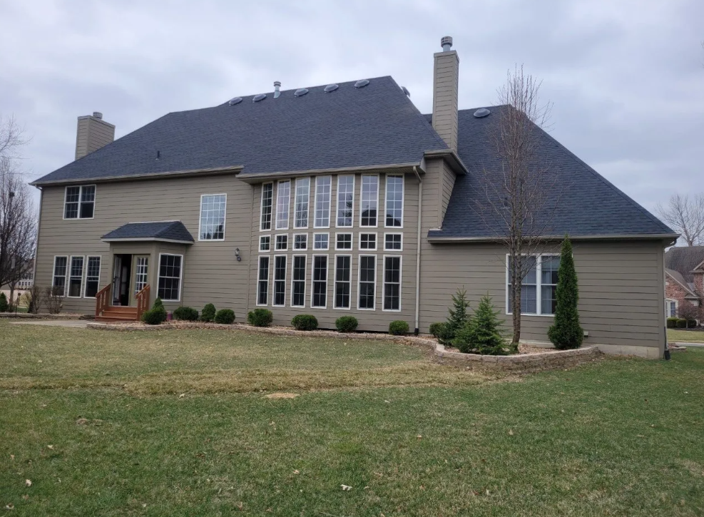 Large beige suburban house with dark roof, tall windows, and a lawn under an overcast sky