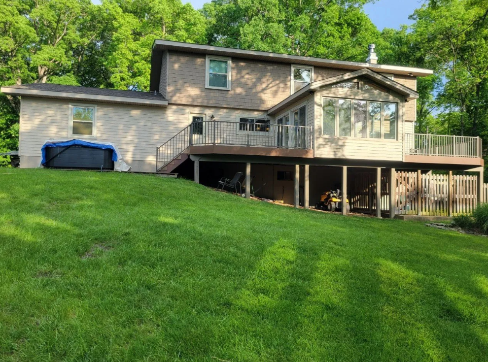 House with a wooden deck on a grassy hillside, surrounded by trees and a blue hot tub.