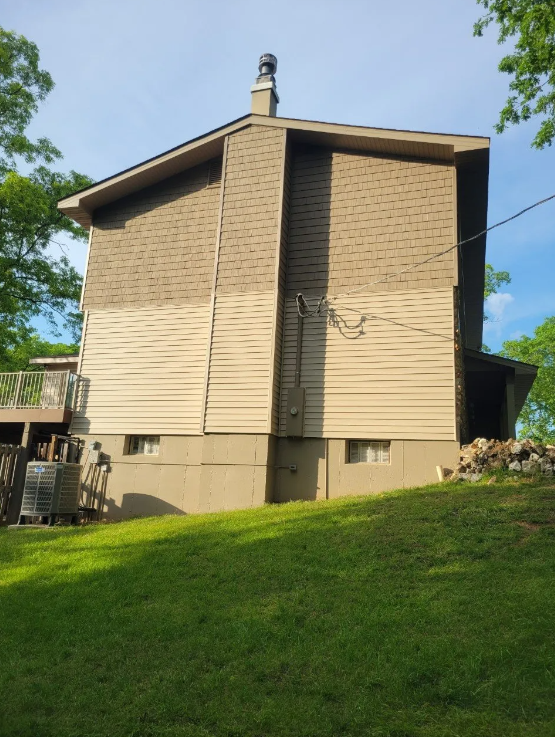 Brown house on a grassy hill with beige siding and a brick chimney under a blue sky.