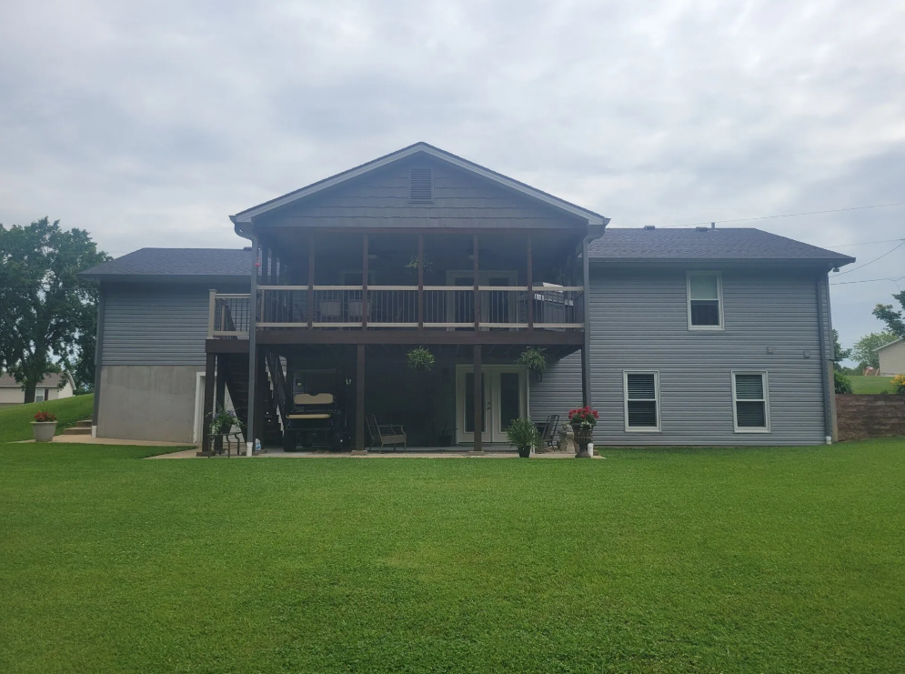Gray two-story house with screened porch and large green lawn under a cloudy sky