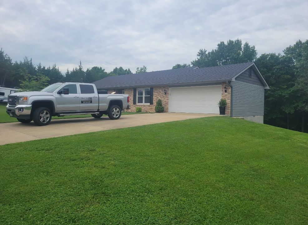 Silver pickup truck parked in a driveway beside a brick house with a large green lawn and cloudy sky.