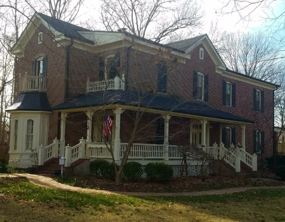 Large brick Victorian house with a wraparound porch and white columns, surrounded by bare trees and lawn.