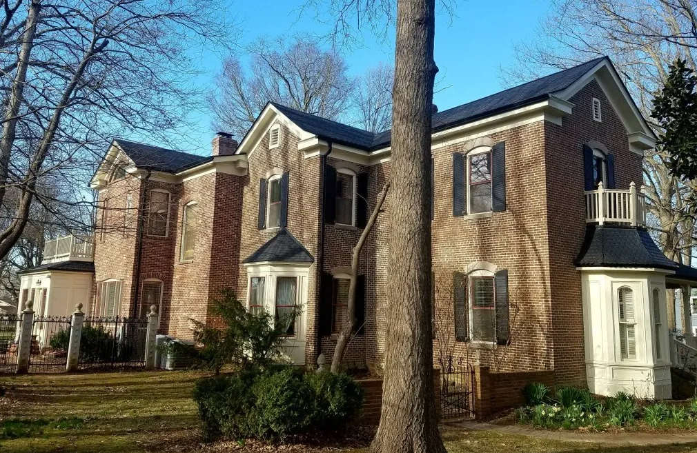 Brick two-story house with white trim, front porch, and trees in the yard under a blue sky