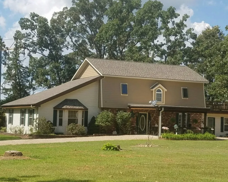 Two-story tan house with a covered porch, white lower wing, and large green lawn under trees