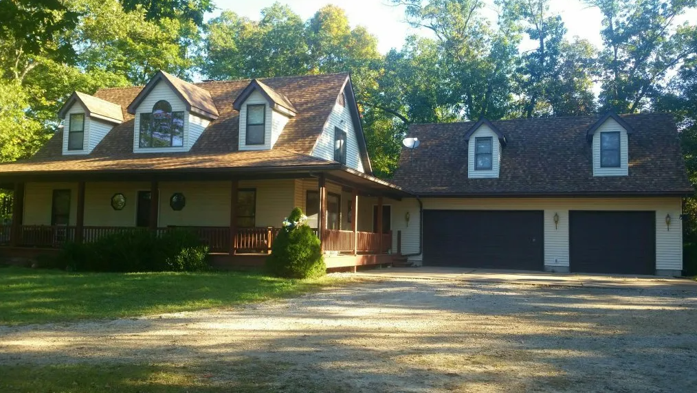 Large house with a covered porch, dormer windows, and a two-car garage surrounded by trees and a gravel driveway
