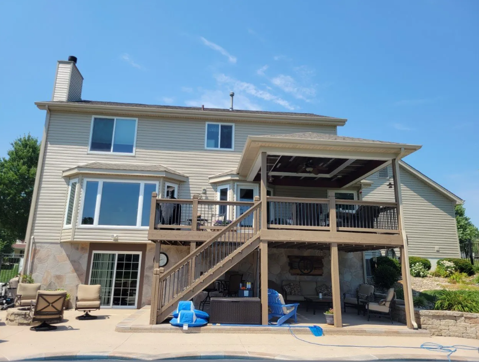 Two-story beige house with a raised wooden deck, stairs, and patio furniture under a blue sky