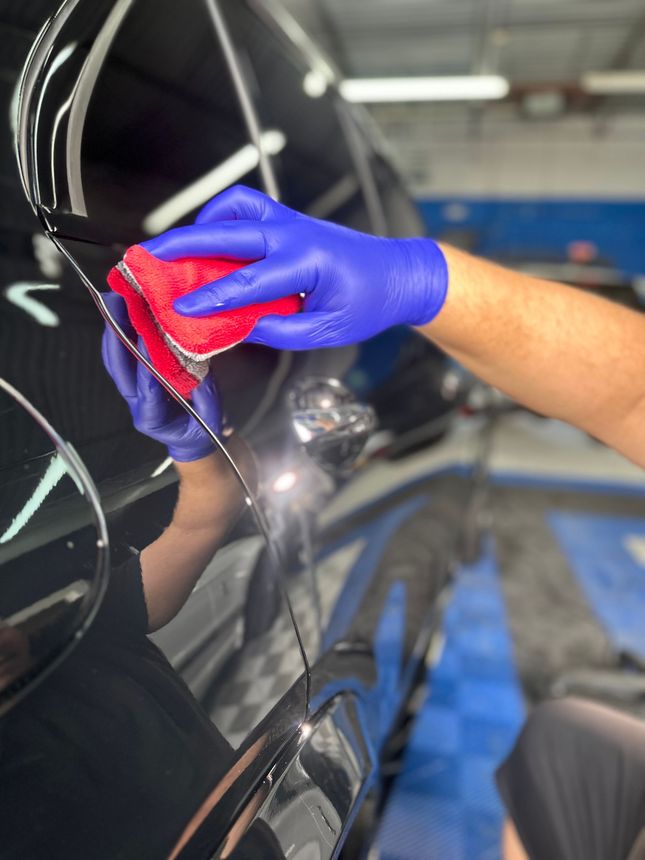 Gloved hand polishing a black car door with a red cloth in a garage