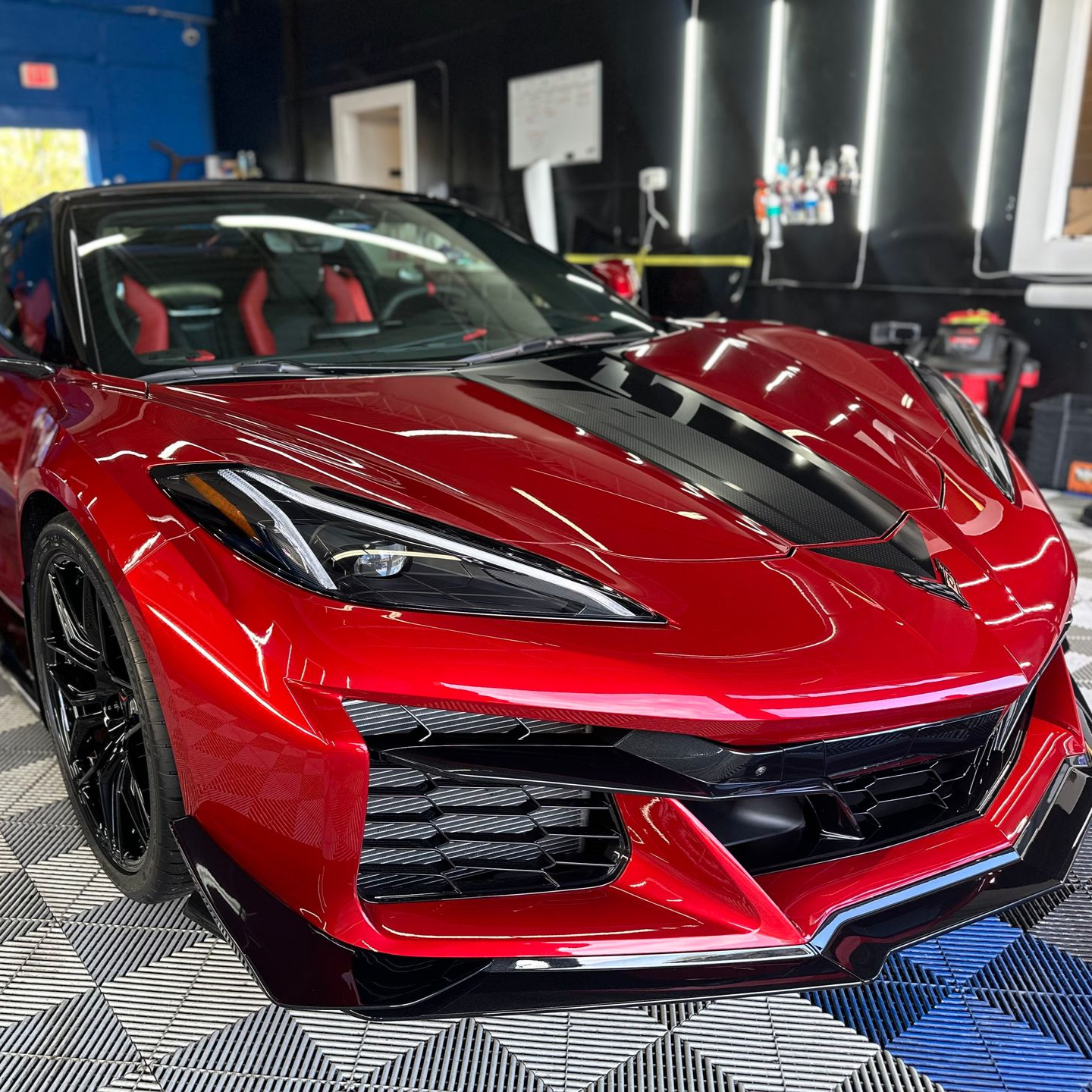 Red sports car with black racing stripes displayed indoors on a checkered floor.