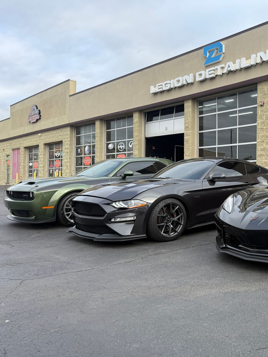 Three sports cars parked outside a detailing shop, including a green muscle car and a black coupe.