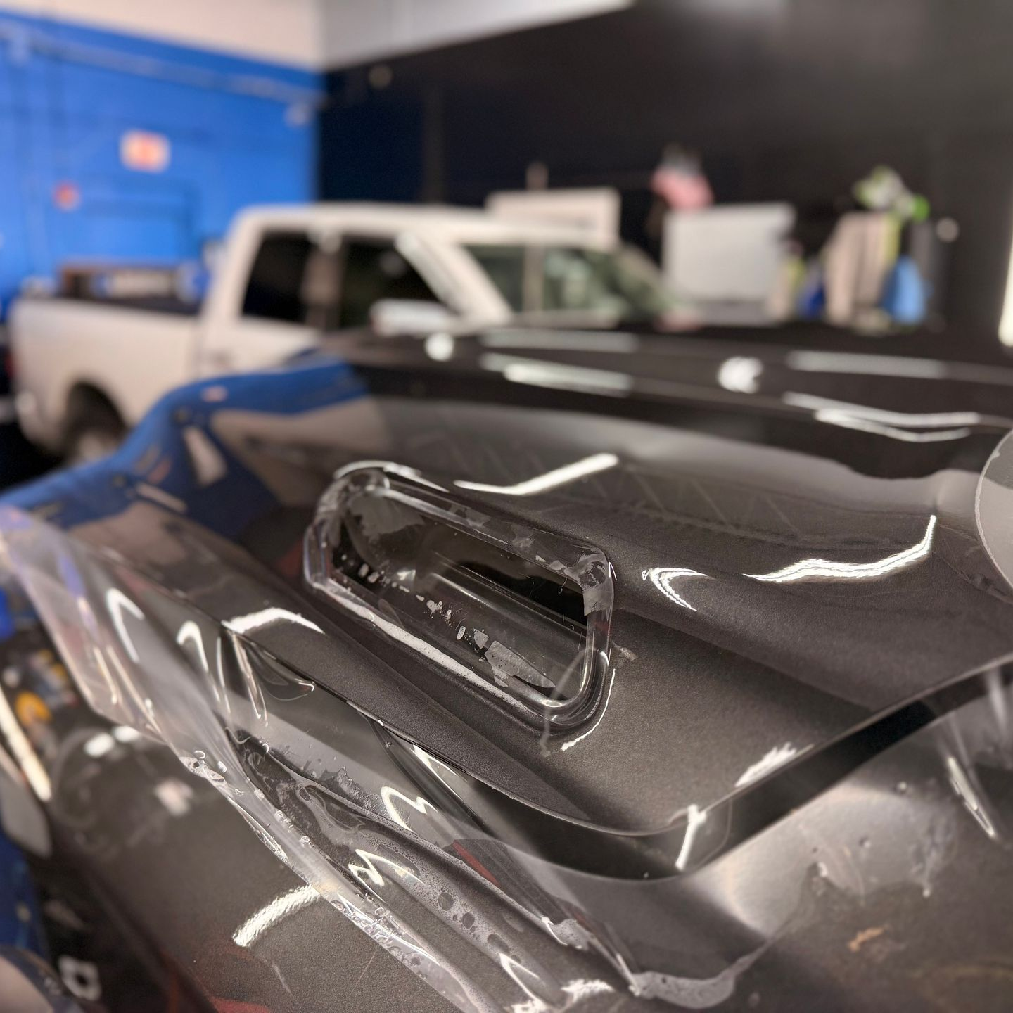 Close-up of a black car hood scoop in a garage, with a white pickup truck in the background