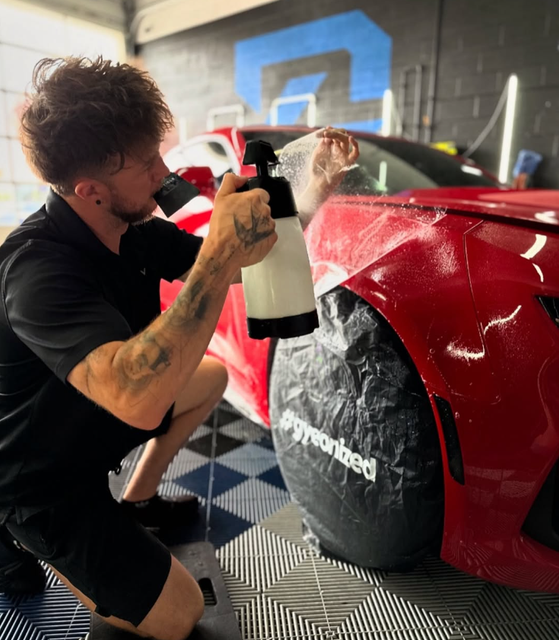 Person sprays a red sports car with a foam bottle in a garage