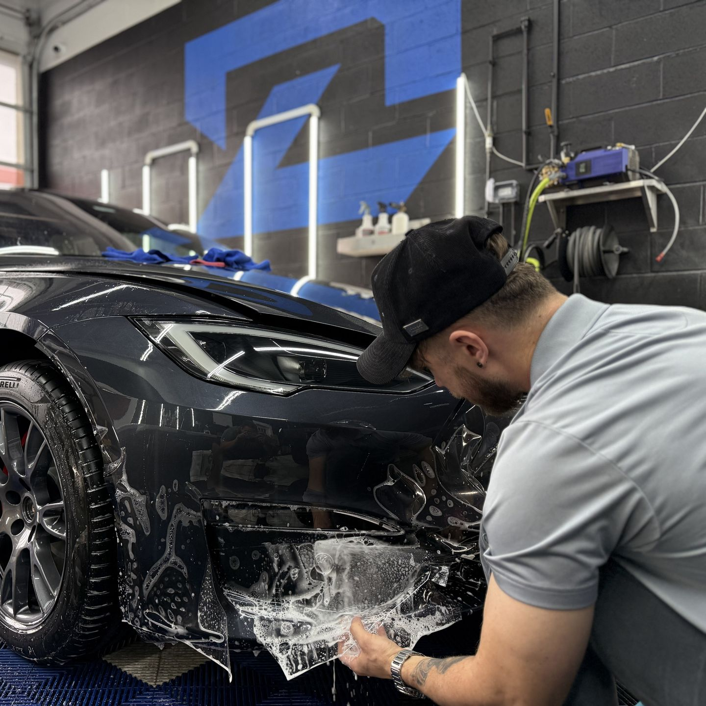 Detailing worker washing a black car in a garage, with soap suds on the front bumper.
