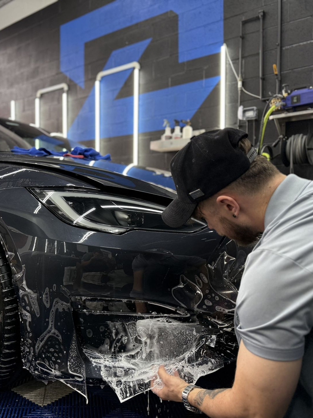 Man in black cap polishing the front bumper of a black car in a garage