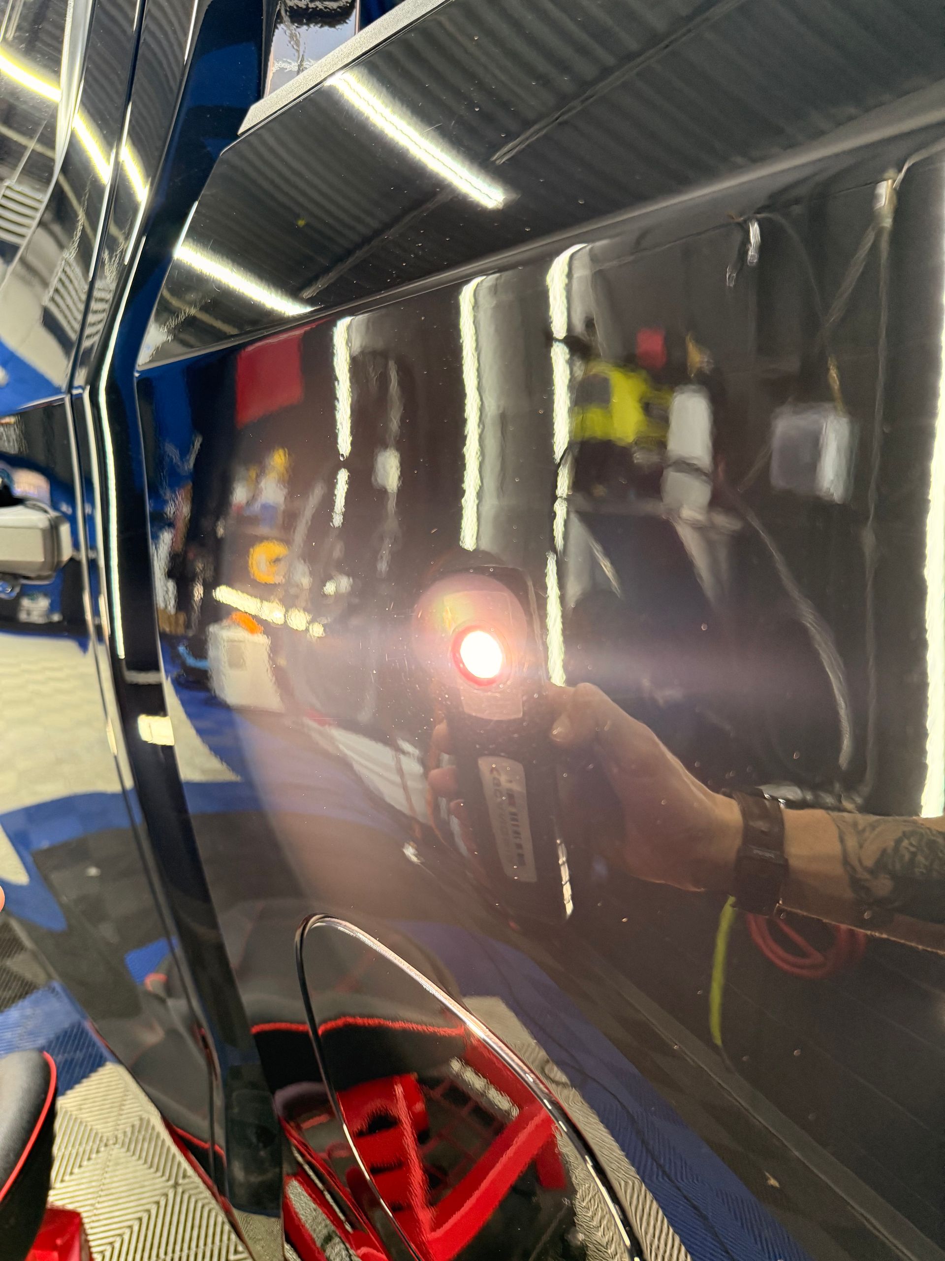 Shiny black car panel in a garage, reflecting a person holding a camera and a bright light.