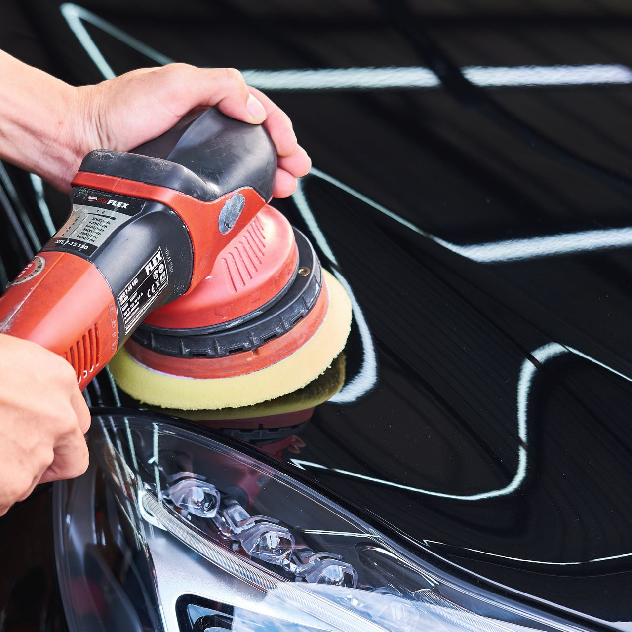 Hands polishing a black car with a red and yellow buffer near the headlight