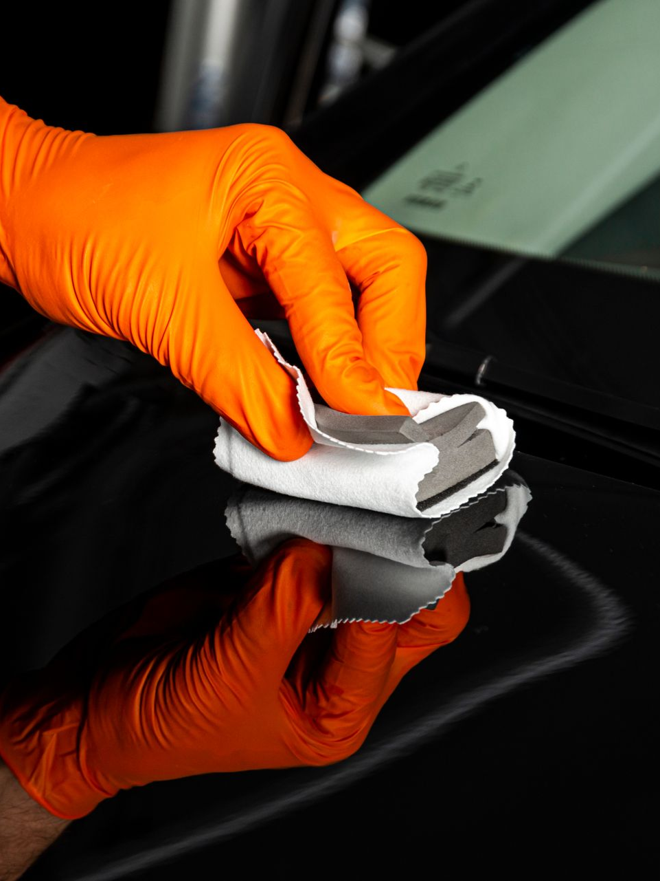 Orange-gloved hand wiping a black car surface with a white cloth.