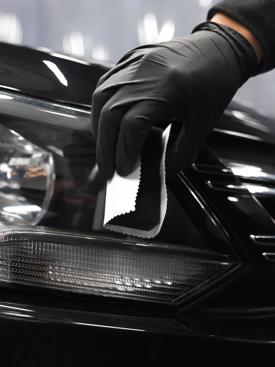 Black-gloved hand wiping a car headlight with a cloth in a glossy garage setting