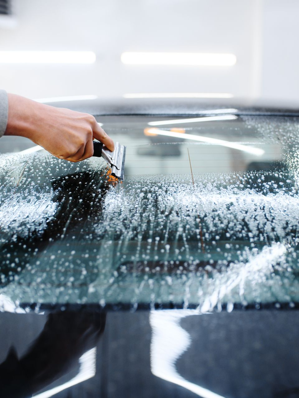 Hand cleaning a car windshield with a sponge, covering it with white soap suds