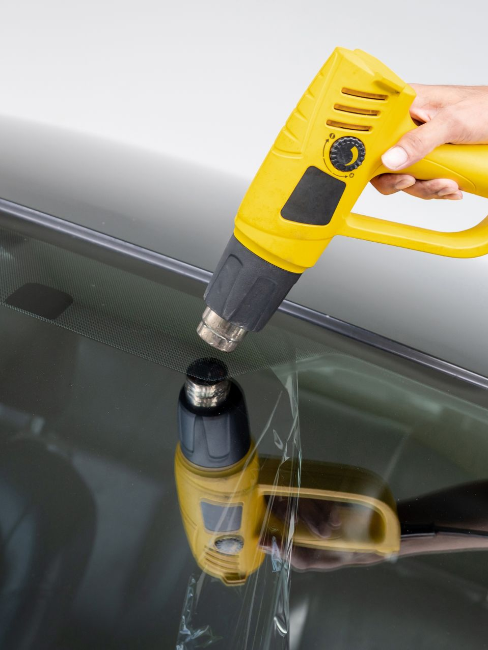 Yellow heat gun aimed at a suction cup on a car windshield, reflecting on the glass