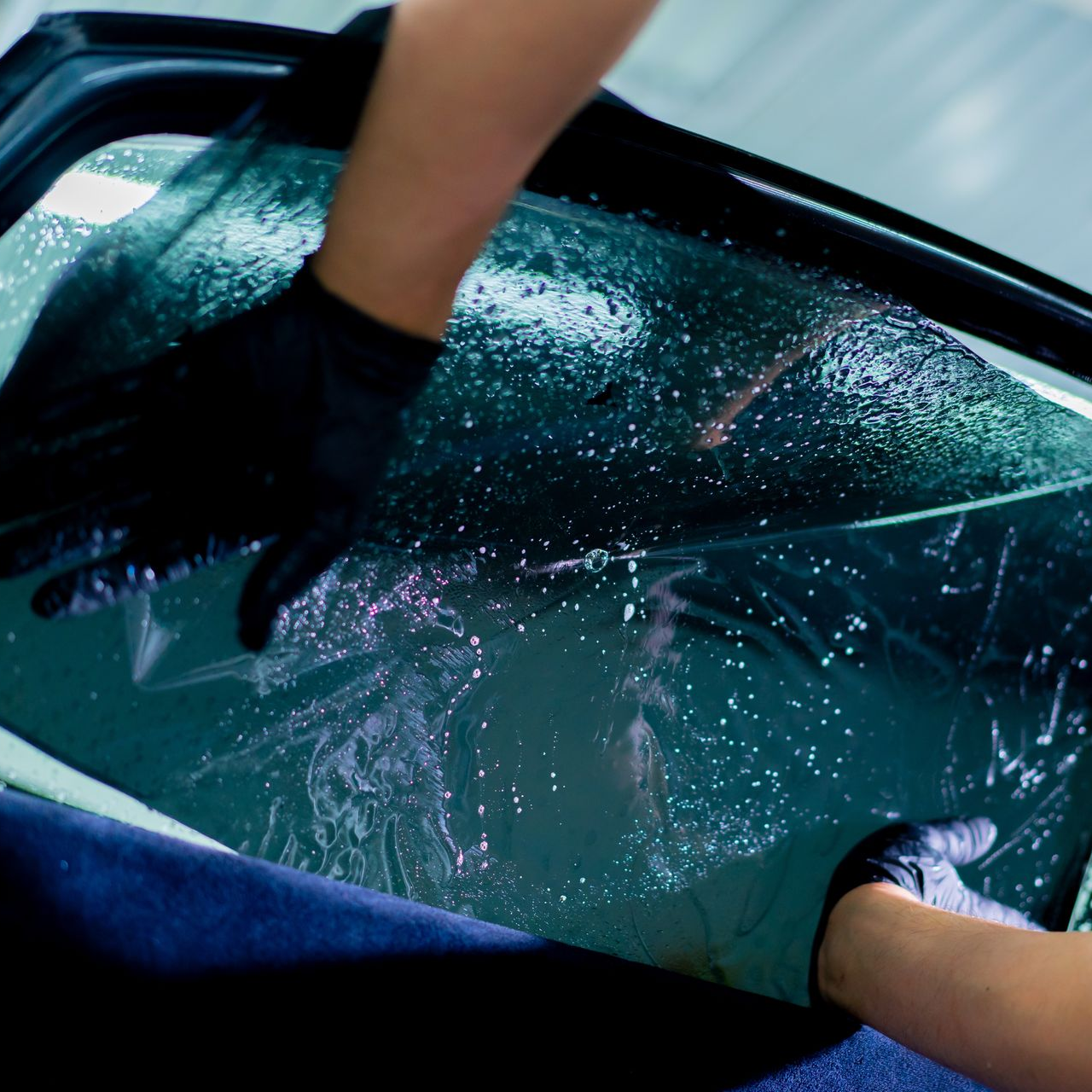 Gloved hands pressing a wet windshield with soapy water and foam during car washing