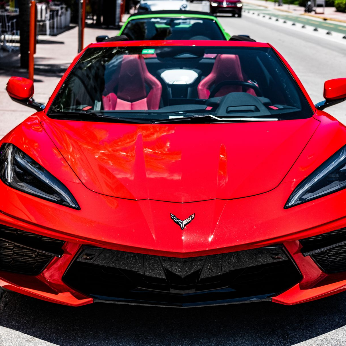 Red Corvette convertible driving on a city street, viewed from the front.
