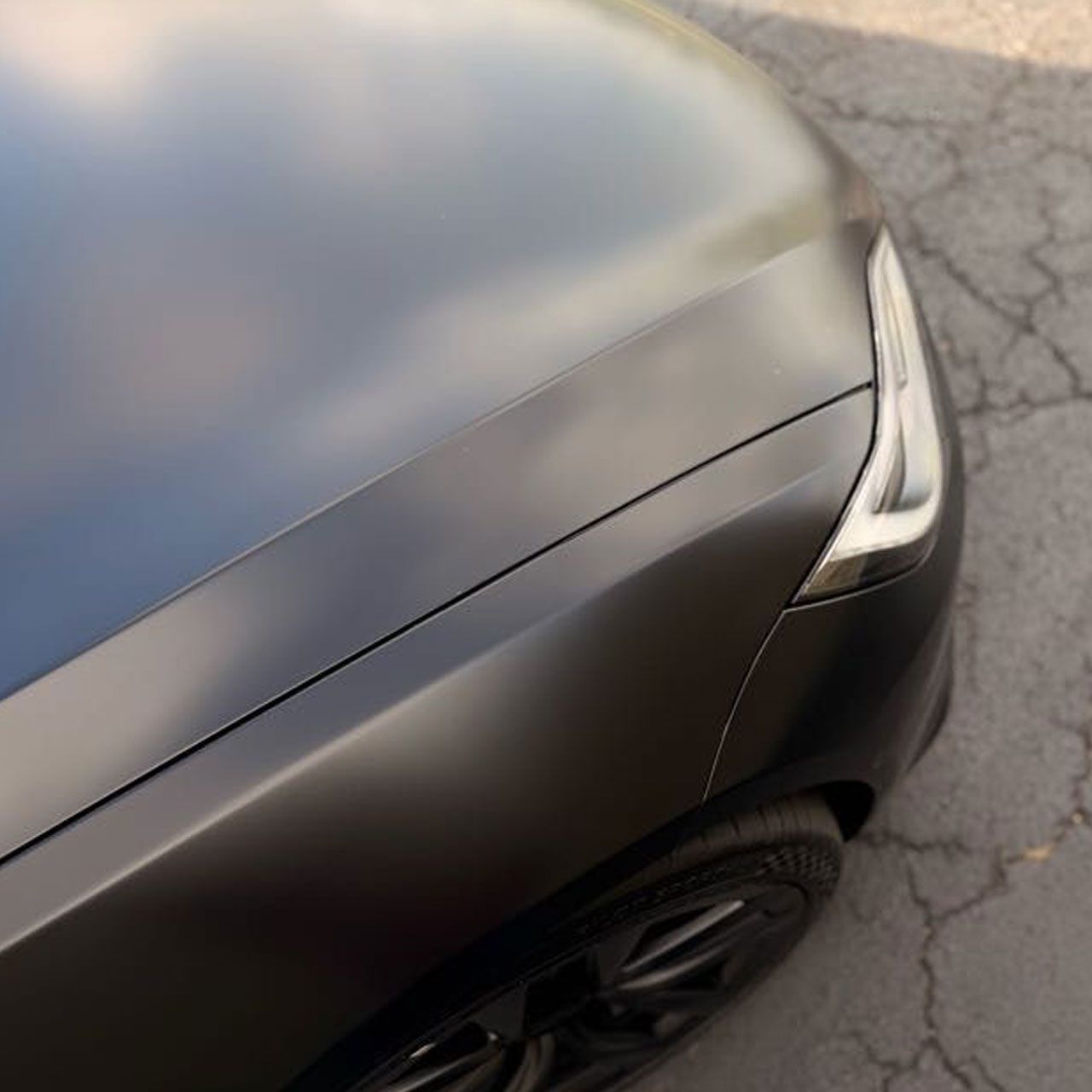 Black car front corner with a cracked headlight and damaged bumper on cracked pavement