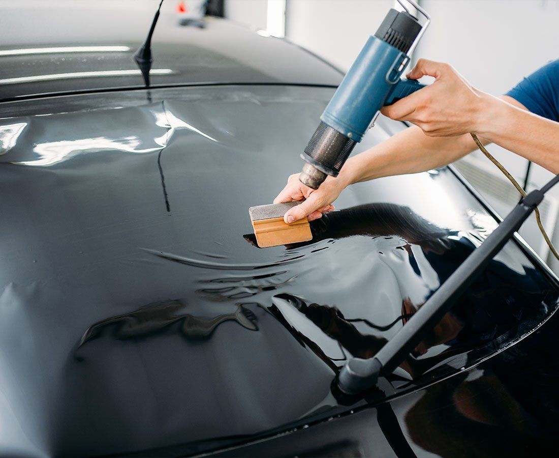 Hand applying black vinyl wrap to a car hood with a heat gun, smoothing bubbles with a squeegee