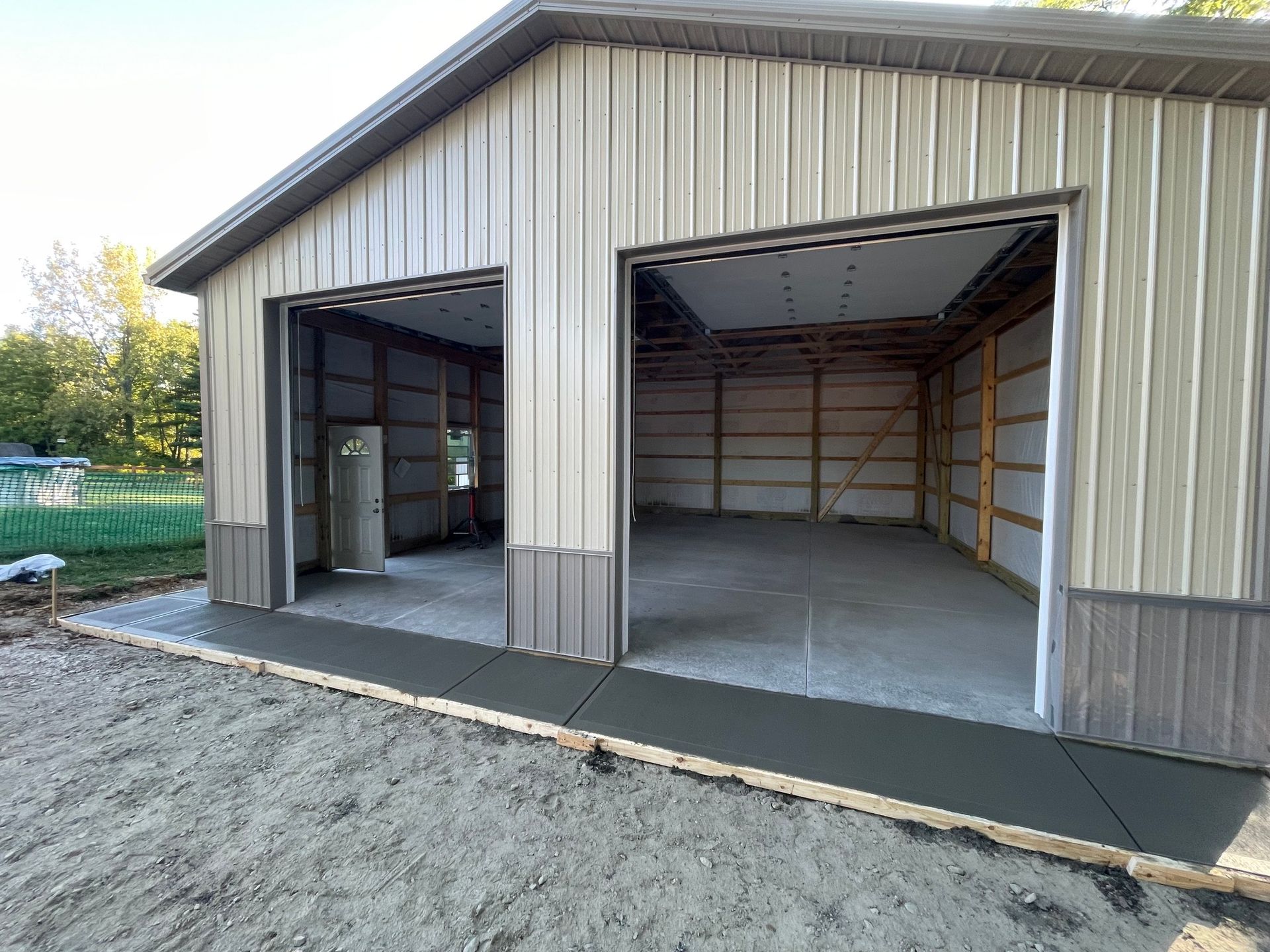 Open steel garage with two wide door openings and a concrete floor, viewed from a gravel driveway.