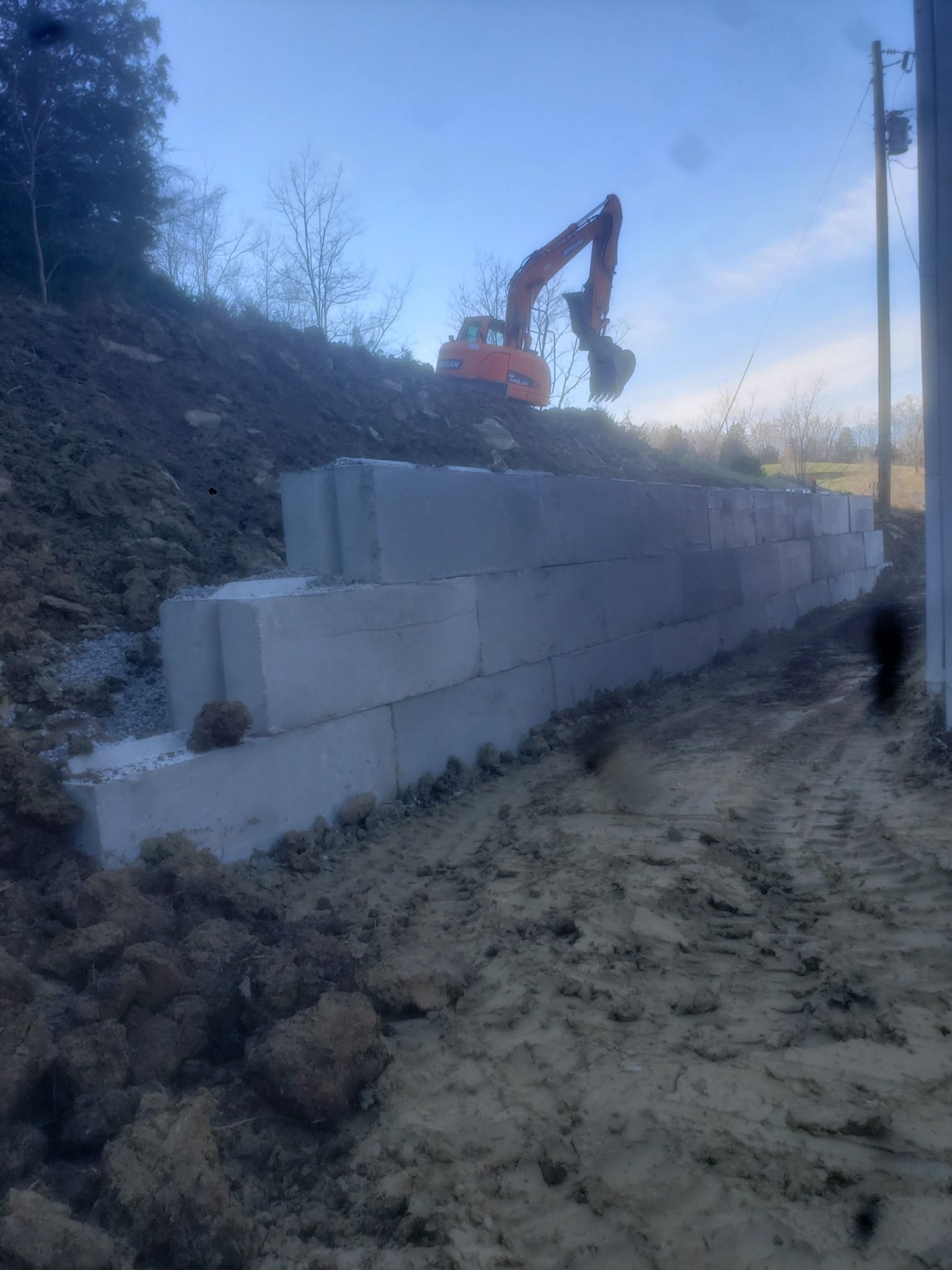 Excavator behind stacked gray concrete blocks on a dirt slope at dusk