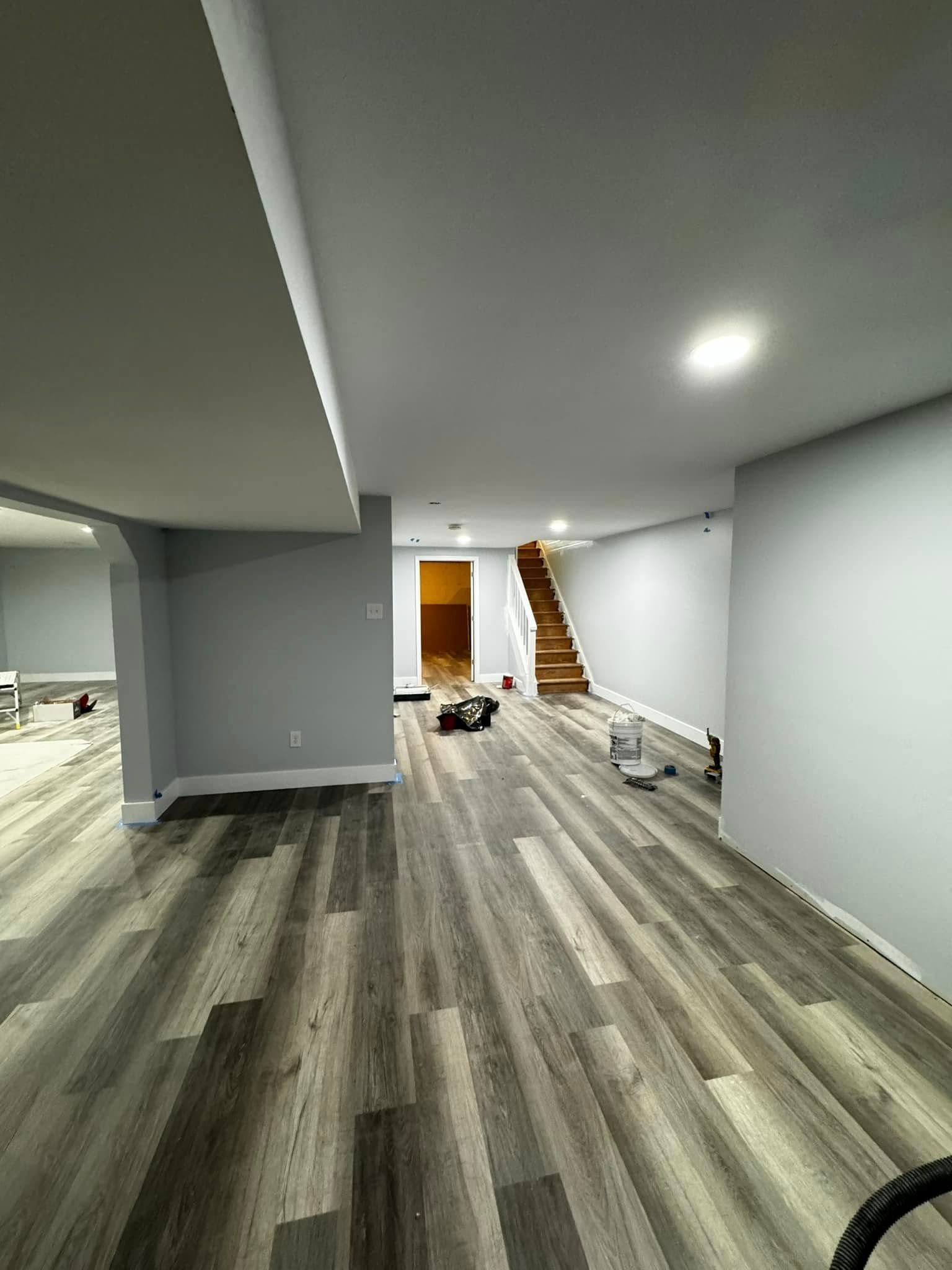 Empty basement room with gray wood floors, white walls, recessed lights, and a staircase in back.