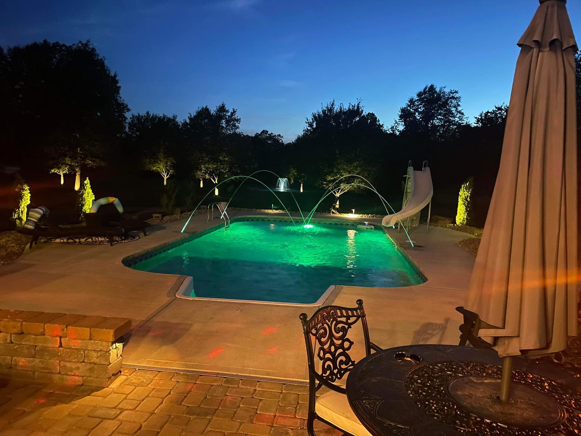 Backyard pool glowing green at night with patio furniture and closed umbrellas under dim lights