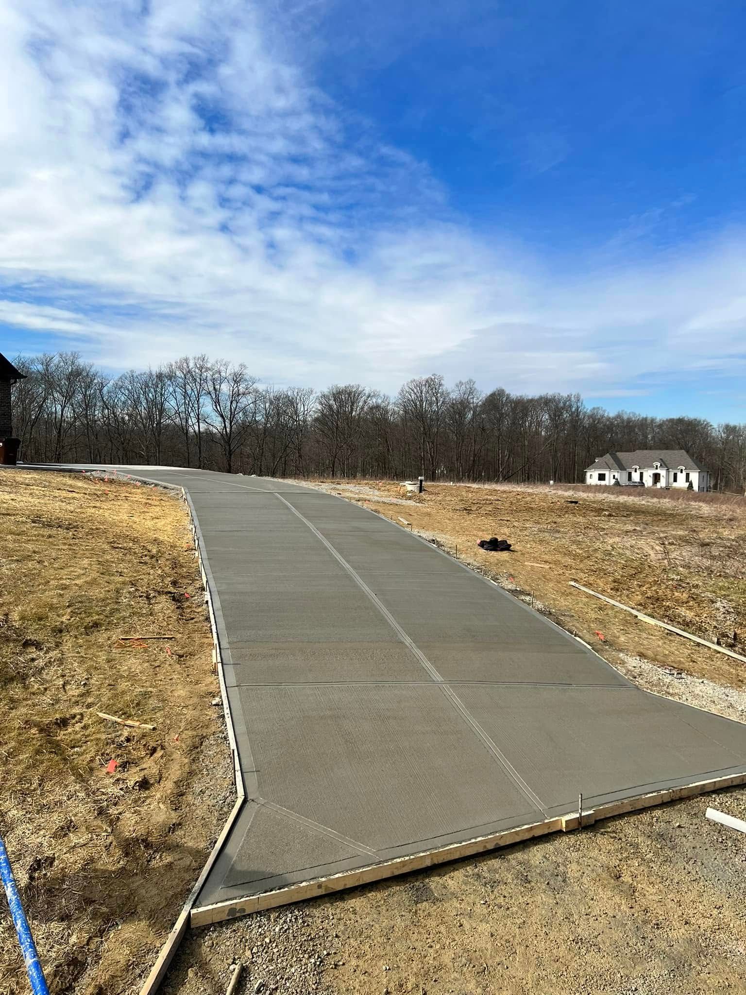 Concrete path through dry field under blue sky, with trees and a house in the distance