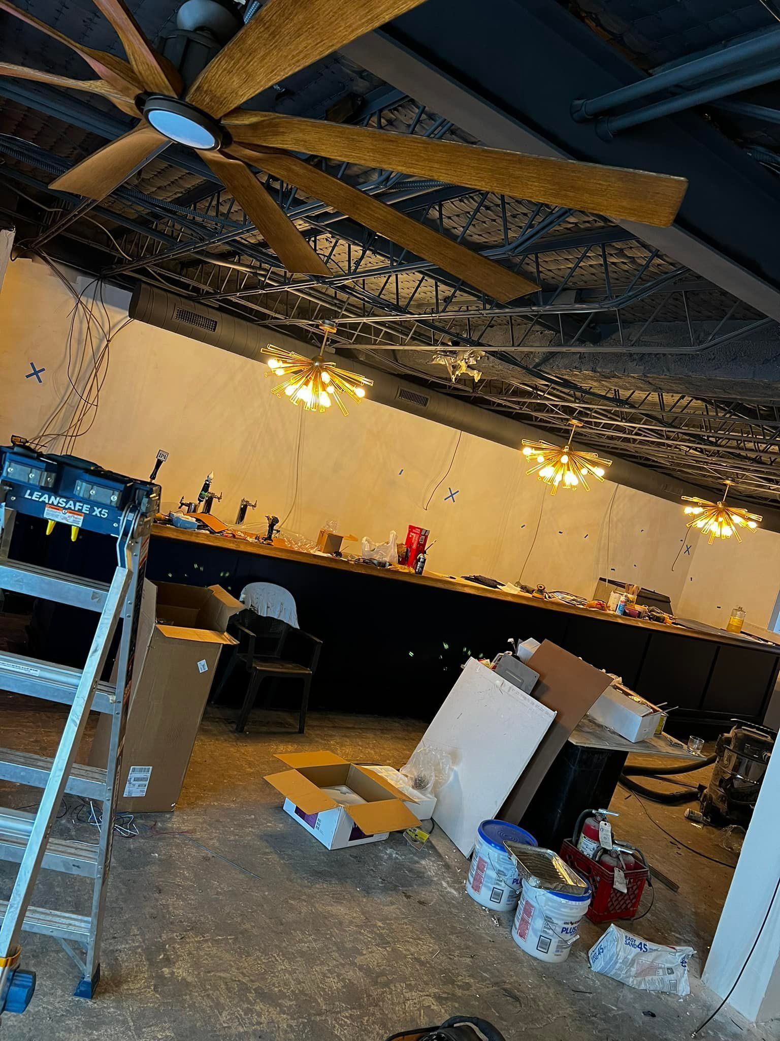 Messy bar area with counter, stools, hanging lights, ceiling fan, and scattered boxes and supplies