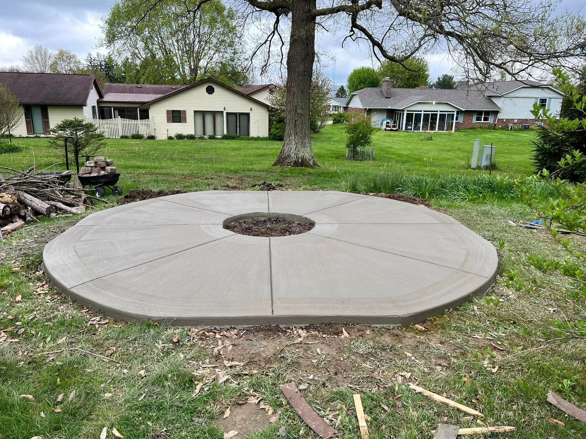 Circular concrete patio in a grassy yard with a tree at the center, houses in the background.