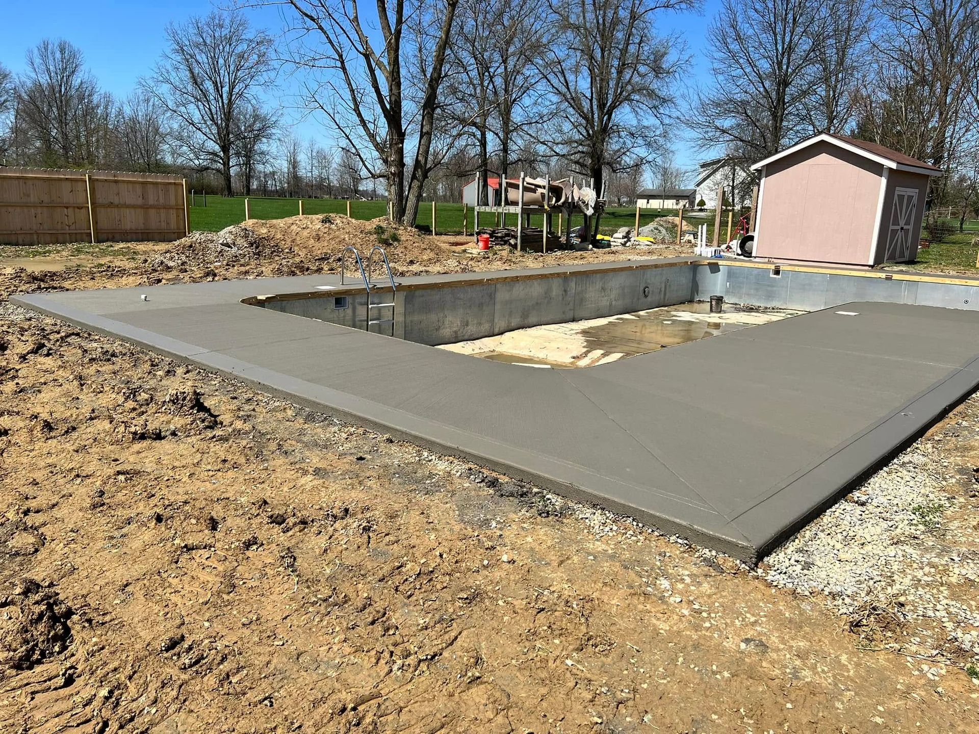 Concrete pool foundation under construction in a backyard, with a pink shed and trees in the background