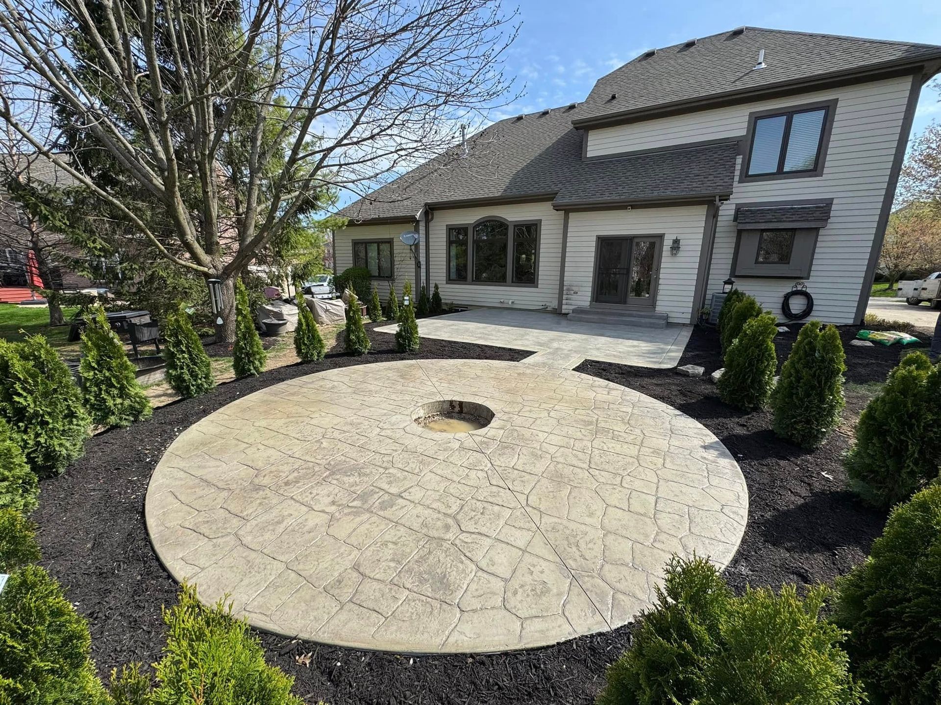 Front yard with a large circular stone patio, bordered by mulch and shrubs in front of a house.