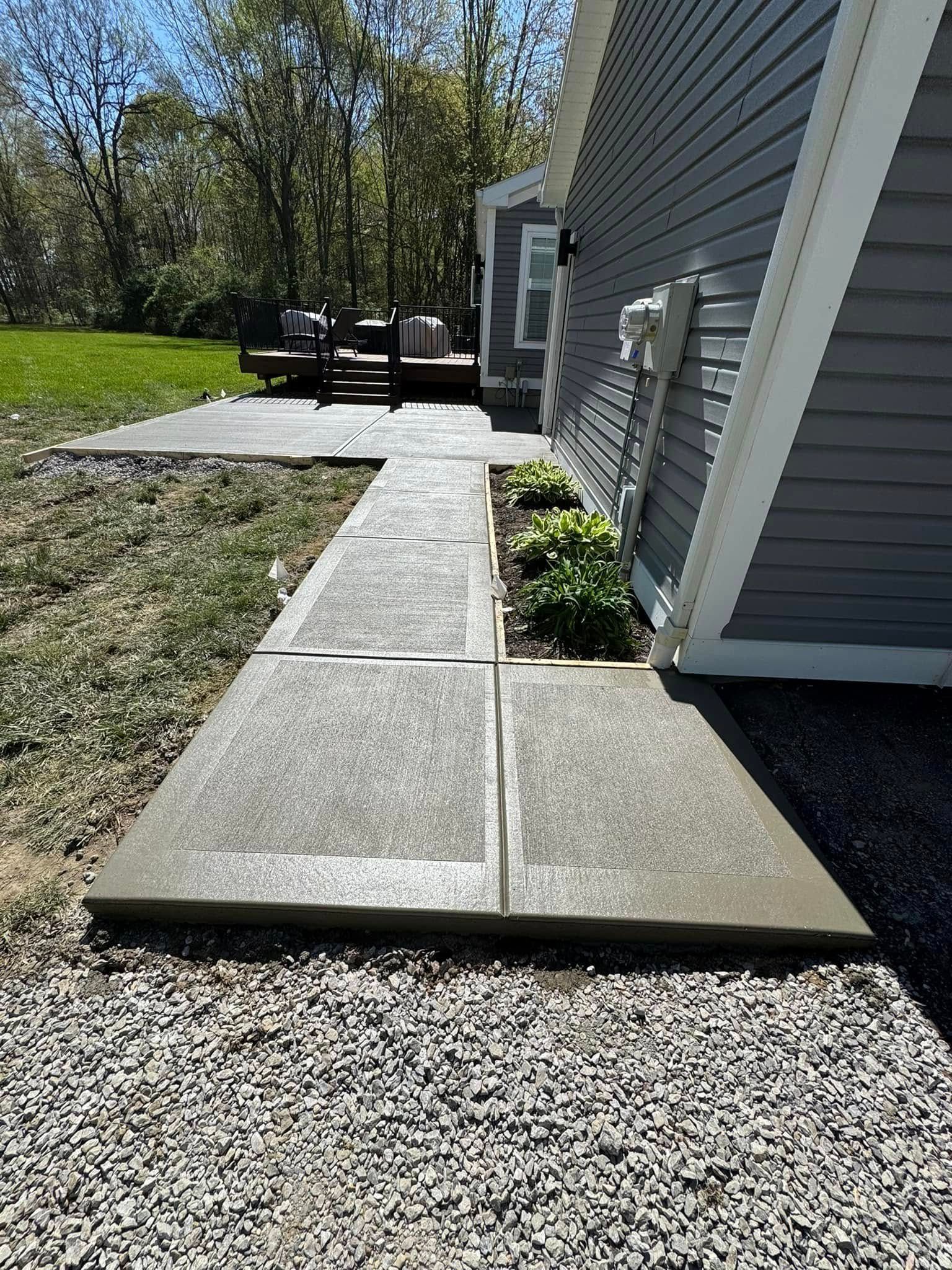 Concrete walkway beside gray house with gravel border and patio in a sunny yard